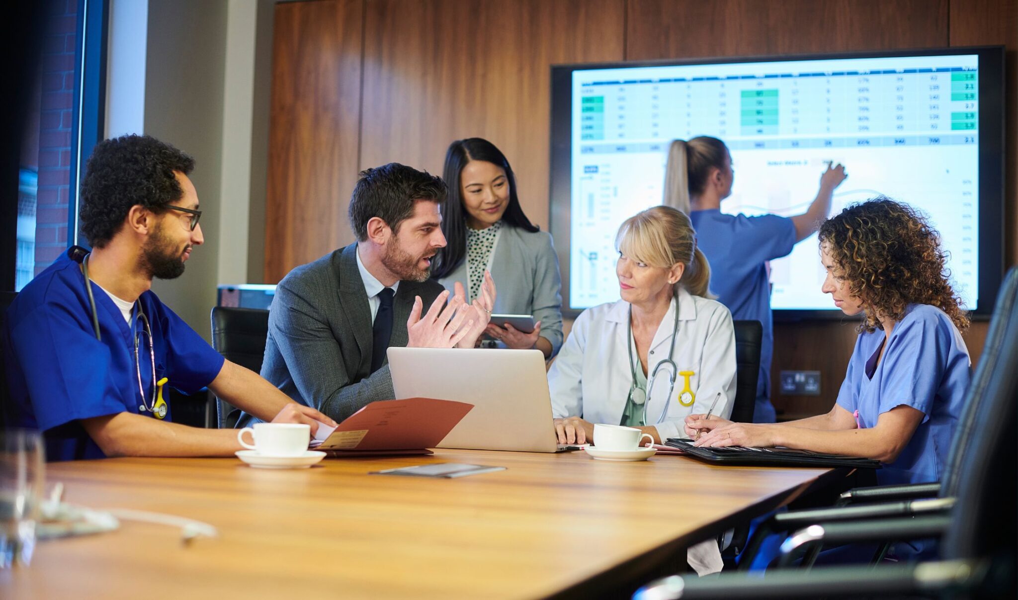 Mixed group of health professions in discussion around a boardroom table. A projected chart of data is being displayed on a screen behind the medical professionsal